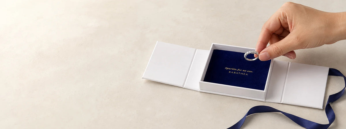 A hand placing a seven-stone silver ring into a partially open Karathea jewelry box with navy ribbons.